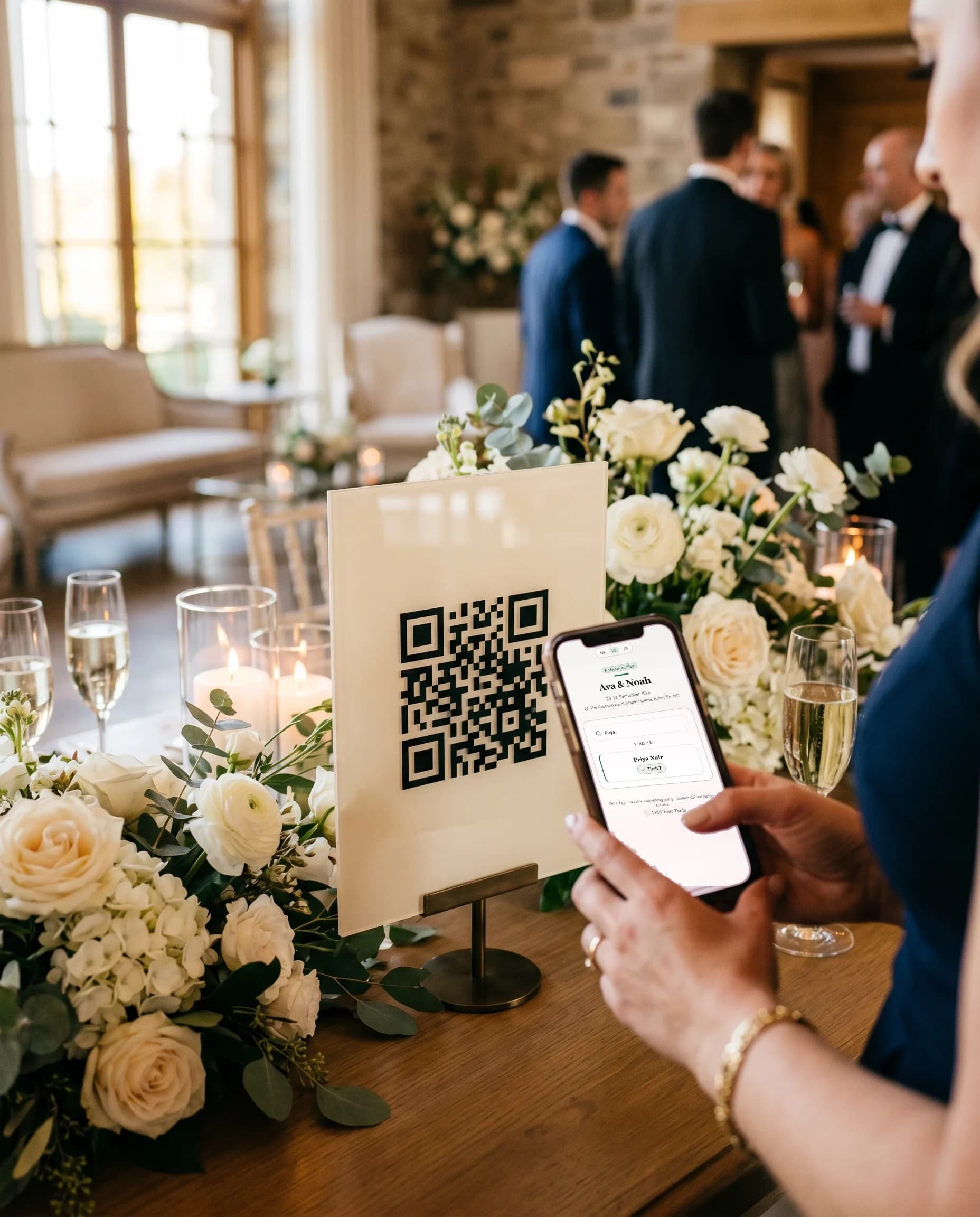Ein Hochzeitsgast scannt mit dem Smartphone den QR-Code auf einem cremefarbenen Willkommensschild; auf dem Display steht der gefundene Sitzplatz — Priya Nair, Tisch 7 — umgeben von weißen Rosen, Kerzen und Champagner bei einer Feier im Kerzenschein